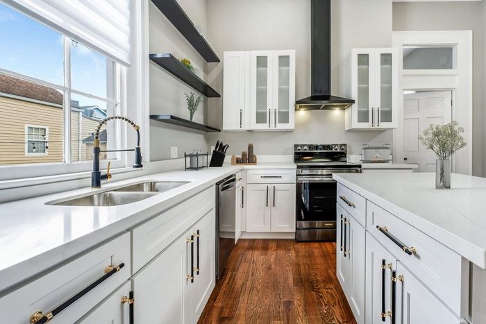 Modern white kitchen with wooden floors, stainless steel appliances, and a kitchen island. A large window provides natural light.